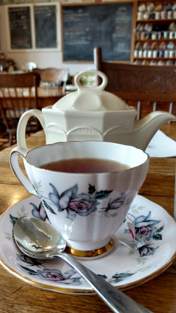 floral teacup, with a white pot in the background. Further back, an older looking tearoom, with wood furniture, a chalkboard, and mugs. 